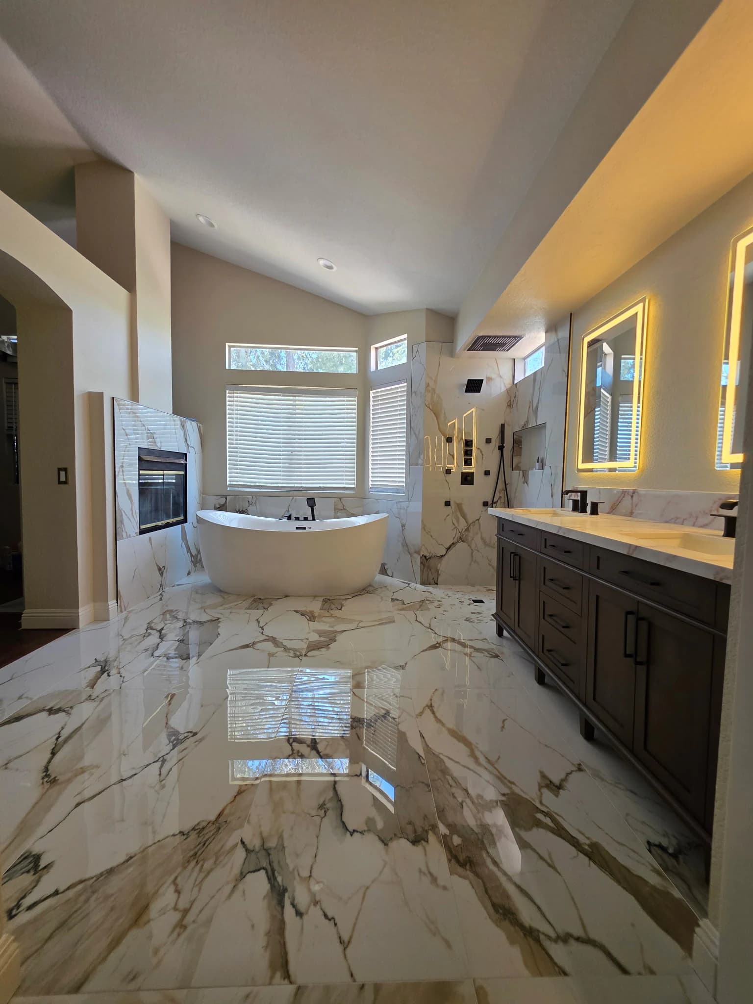 Completed marble-look bathroom viewed across the tiled floor toward the tub, shower, fireplace, and double vanity